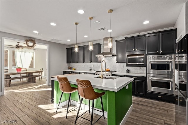 kitchen with pendant lighting, wall chimney exhaust hood, stainless steel appliances, a kitchen breakfast bar, and a center island with sink