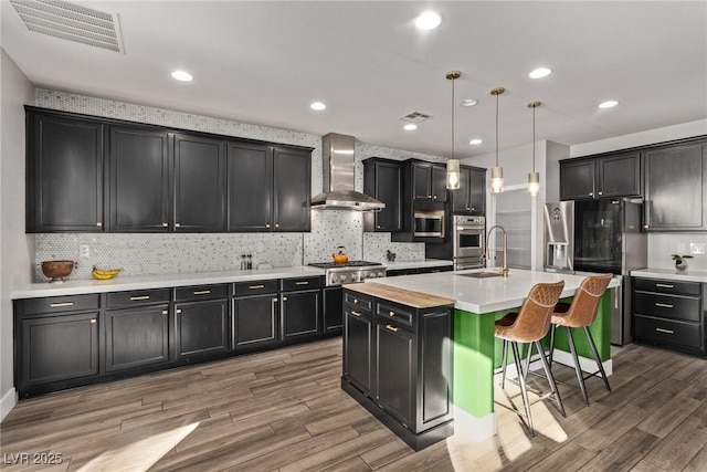 kitchen featuring pendant lighting, appliances with stainless steel finishes, a kitchen island with sink, a breakfast bar, and wall chimney range hood