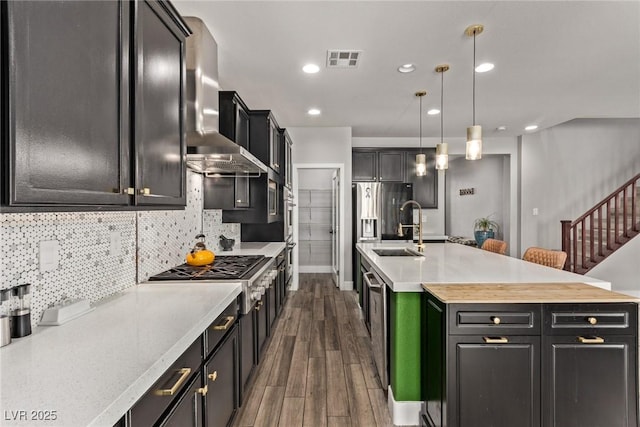 kitchen featuring decorative light fixtures, wall chimney exhaust hood, sink, an island with sink, and butcher block counters