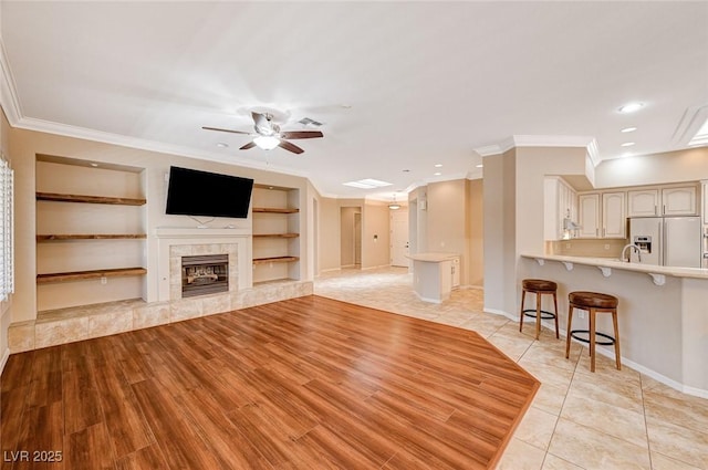 unfurnished living room with sink, built in features, ceiling fan, a tile fireplace, and crown molding