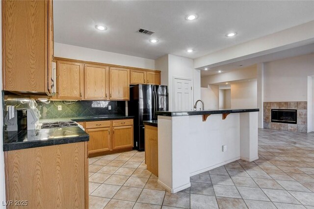 kitchen with black refrigerator, backsplash, light tile patterned floors, a tile fireplace, and an island with sink