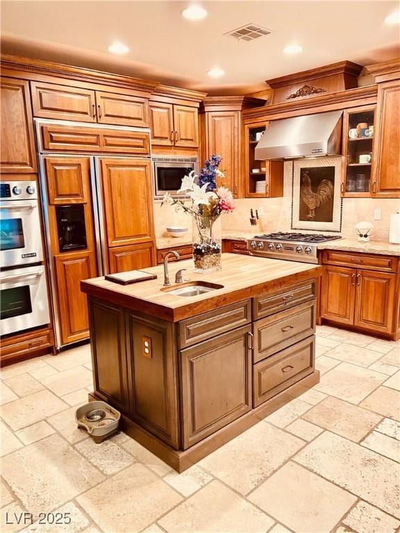 kitchen featuring sink, wooden counters, stainless steel appliances, a kitchen island with sink, and wall chimney range hood