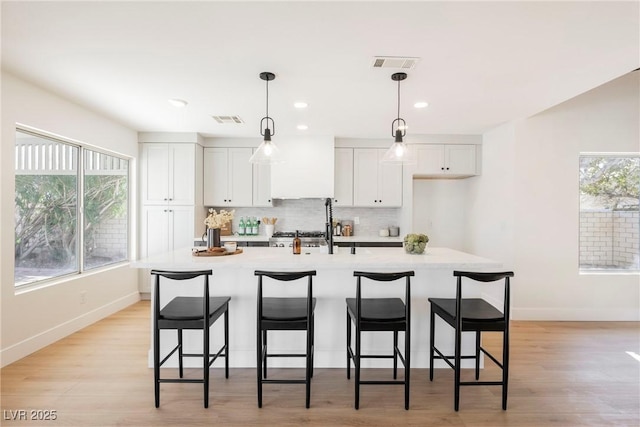 kitchen featuring pendant lighting, tasteful backsplash, white cabinetry, a kitchen island with sink, and light hardwood / wood-style flooring