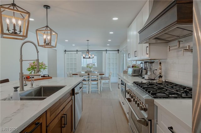 kitchen featuring custom exhaust hood, appliances with stainless steel finishes, white cabinetry, hanging light fixtures, and a barn door