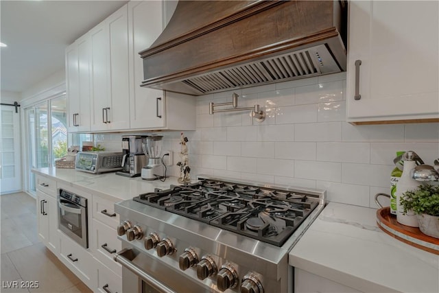 kitchen featuring light stone countertops, appliances with stainless steel finishes, white cabinetry, custom range hood, and decorative backsplash