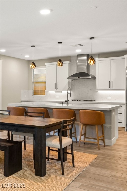 kitchen featuring hanging light fixtures, light wood-type flooring, white cabinetry, and wall chimney range hood