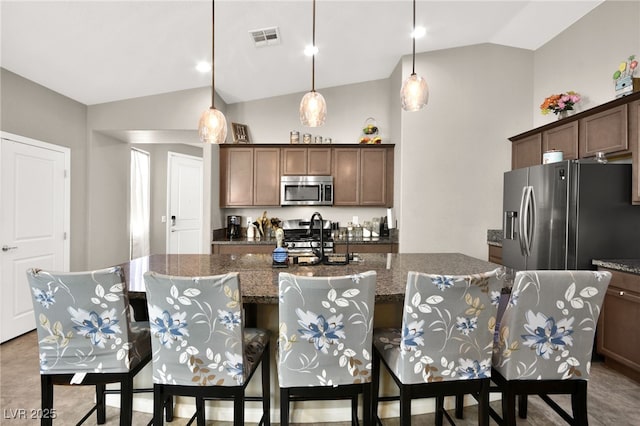 kitchen featuring dark stone counters, decorative light fixtures, stainless steel appliances, a center island with sink, and lofted ceiling