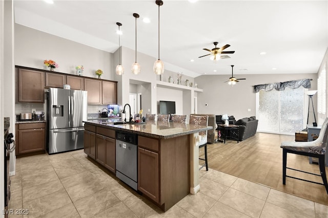 kitchen featuring light tile patterned floors, an island with sink, appliances with stainless steel finishes, dark stone countertops, and decorative light fixtures