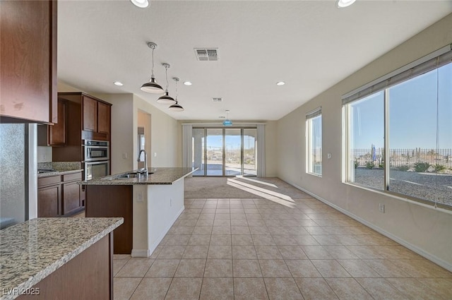 kitchen with sink, double oven, an island with sink, pendant lighting, and light stone countertops