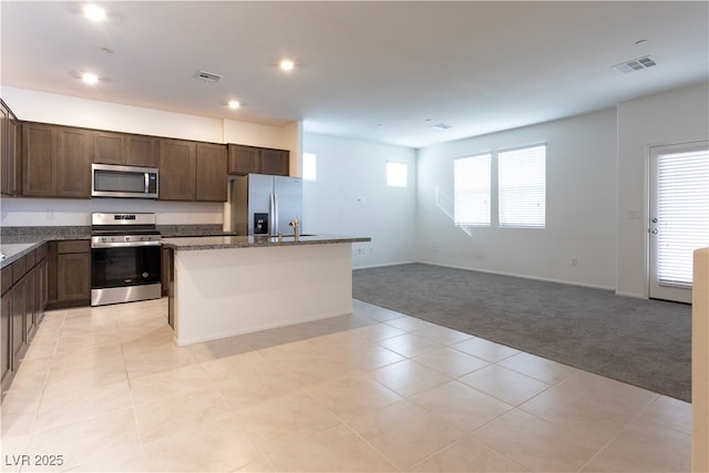 kitchen with appliances with stainless steel finishes, dark brown cabinetry, an island with sink, light colored carpet, and dark stone counters