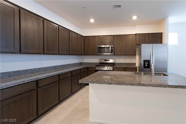 kitchen featuring an island with sink, appliances with stainless steel finishes, dark stone countertops, and dark brown cabinetry