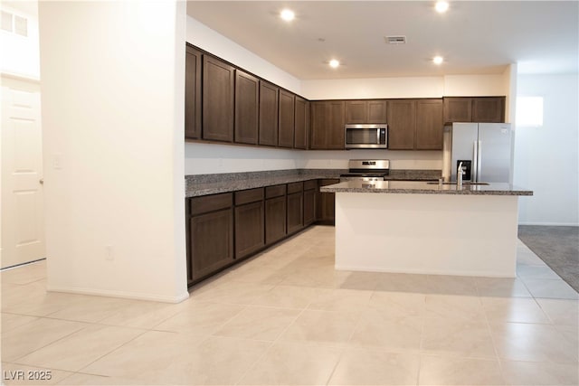 kitchen featuring stainless steel appliances, light tile patterned flooring, a kitchen island with sink, and dark brown cabinetry