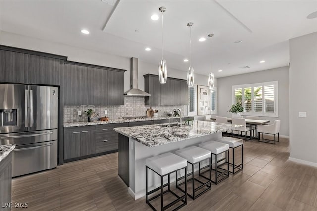 kitchen with a sink, a kitchen breakfast bar, appliances with stainless steel finishes, backsplash, and wall chimney exhaust hood