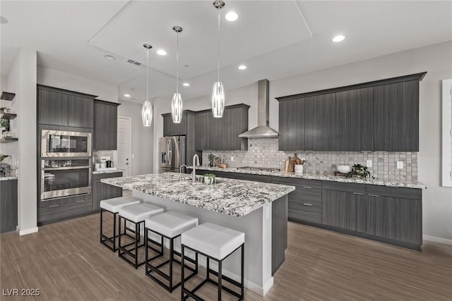 kitchen featuring appliances with stainless steel finishes, decorative light fixtures, sink, a kitchen island with sink, and wall chimney range hood