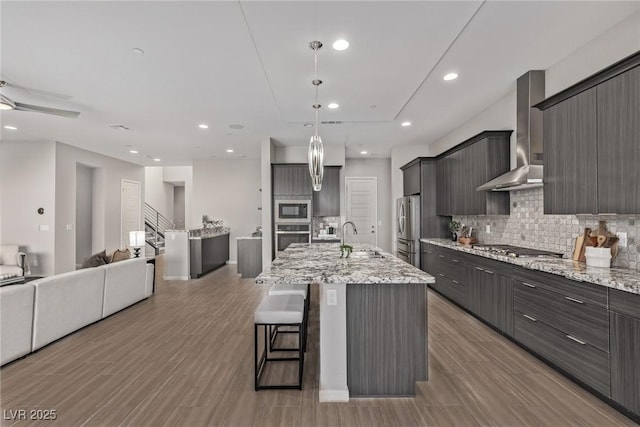 kitchen with wall chimney exhaust hood, open floor plan, stainless steel appliances, a kitchen bar, and a sink