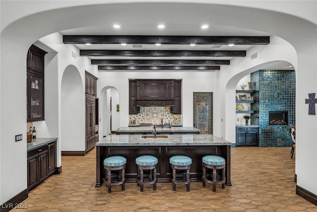 kitchen with dark brown cabinetry, an island with sink, beamed ceiling, a fireplace, and decorative backsplash