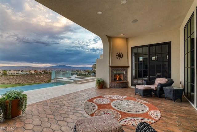 patio terrace at dusk featuring a lit fireplace, a mountain view, and an outdoor pool