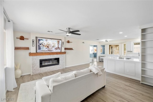 living area featuring visible vents, a ceiling fan, a glass covered fireplace, light wood-type flooring, and baseboards