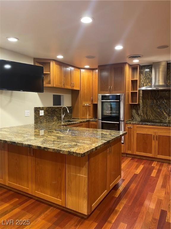 kitchen featuring wall chimney exhaust hood, dark hardwood / wood-style flooring, double oven, stone countertops, and kitchen peninsula