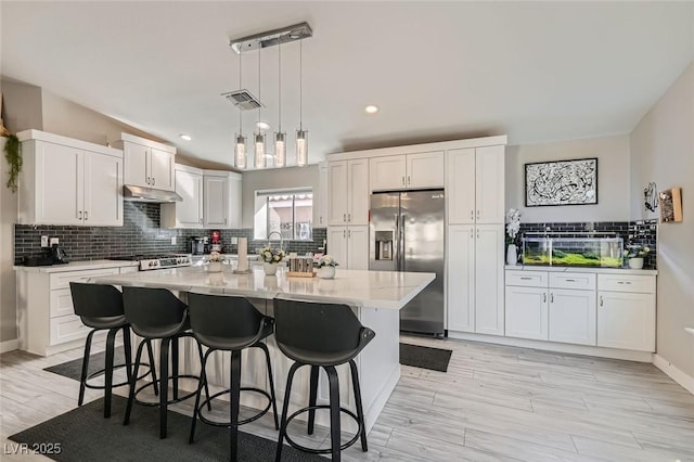 kitchen featuring a kitchen island with sink, backsplash, stainless steel refrigerator with ice dispenser, white cabinets, and decorative light fixtures