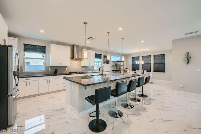 kitchen featuring wall chimney range hood, appliances with stainless steel finishes, dark stone countertops, white cabinets, and a kitchen bar