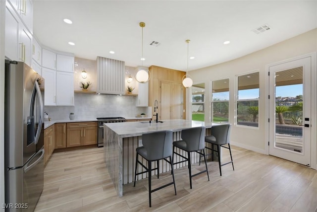 kitchen featuring appliances with stainless steel finishes, pendant lighting, white cabinets, light stone countertops, and a center island with sink