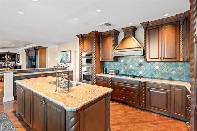 kitchen featuring premium range hood, tasteful backsplash, light stone counters, a center island with sink, and stainless steel double oven