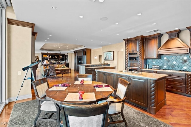 kitchen with hardwood / wood-style flooring, backsplash, light stone counters, custom range hood, and a center island with sink