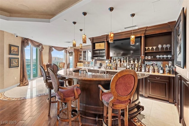 bar featuring dark brown cabinets, light stone counters, decorative light fixtures, a raised ceiling, and light wood-type flooring