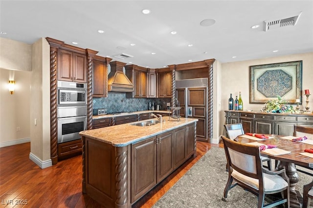 kitchen with dark hardwood / wood-style flooring, custom exhaust hood, a kitchen island with sink, stainless steel appliances, and light stone countertops