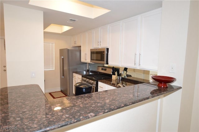 kitchen featuring white cabinetry, a skylight, stainless steel appliances, kitchen peninsula, and dark stone counters