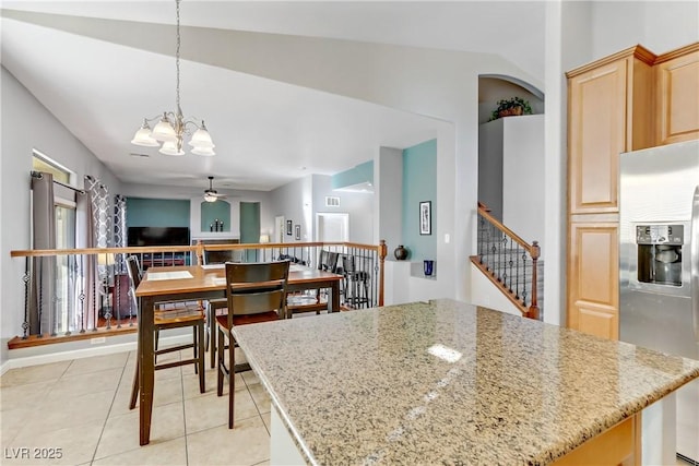 kitchen with stainless steel refrigerator with ice dispenser, light brown cabinetry, light stone counters, pendant lighting, and ceiling fan with notable chandelier