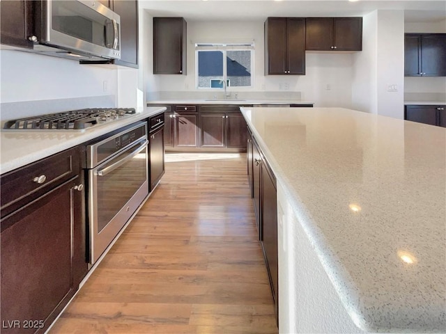 kitchen featuring stainless steel appliances, light wood-style flooring, a sink, dark brown cabinetry, and light stone countertops