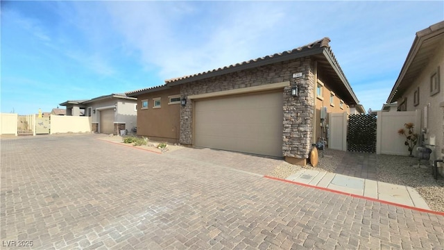 view of property exterior with an attached garage, stone siding, stucco siding, a gate, and decorative driveway