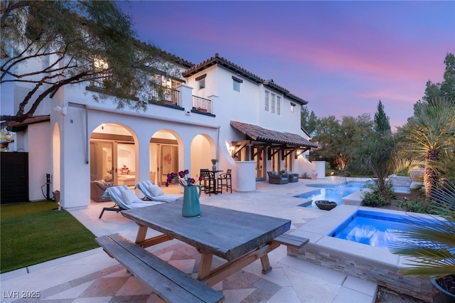 back house at dusk featuring a swimming pool with hot tub, a patio, and a balcony