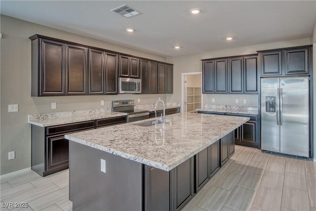 kitchen with appliances with stainless steel finishes, dark brown cabinets, an island with sink, light stone countertops, and sink