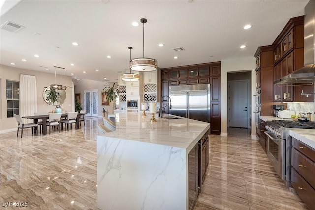 kitchen featuring sink, hanging light fixtures, high end appliances, light stone countertops, and a large island with sink