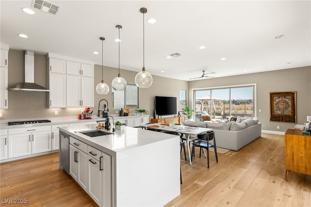 kitchen with pendant lighting, white cabinetry, sink, wall chimney range hood, and a center island with sink