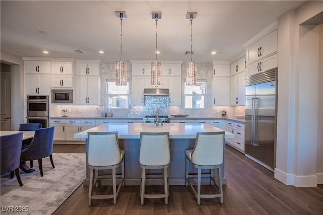 kitchen featuring built in appliances, decorative light fixtures, dark hardwood / wood-style floors, an island with sink, and white cabinets