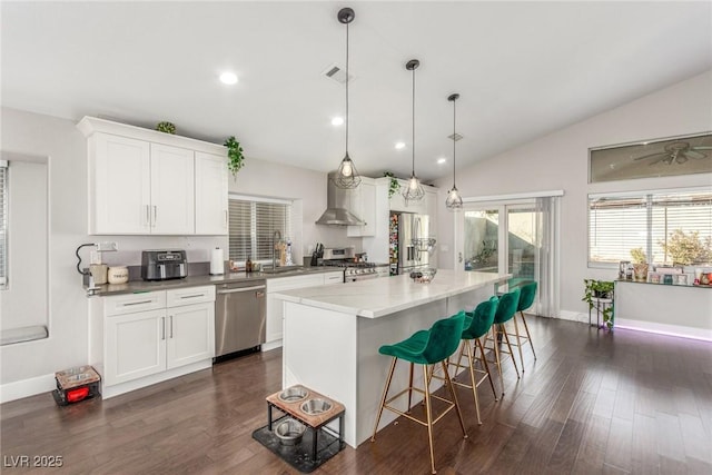 kitchen featuring a kitchen island, appliances with stainless steel finishes, vaulted ceiling, wall chimney range hood, and a kitchen bar