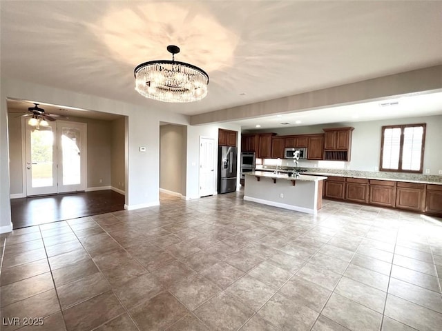 kitchen with ceiling fan with notable chandelier, an island with sink, hanging light fixtures, stainless steel appliances, and light stone countertops