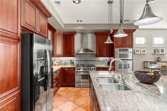 kitchen featuring sink, light stone counters, hanging light fixtures, appliances with stainless steel finishes, and wall chimney range hood