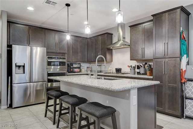 kitchen featuring decorative light fixtures, a center island with sink, dark brown cabinets, stainless steel appliances, and wall chimney range hood