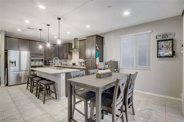 kitchen featuring dark brown cabinets, light tile patterned floors, hanging light fixtures, stainless steel appliances, and wall chimney range hood