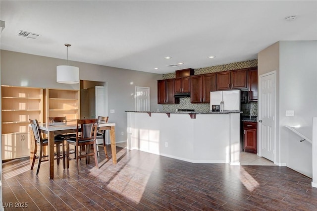 kitchen with dark wood-type flooring, a breakfast bar area, hanging light fixtures, dark stone counters, and backsplash