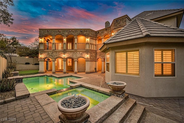 back house at dusk with a balcony, a pool with hot tub, and a patio area