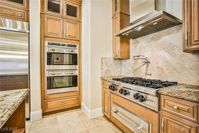 kitchen featuring light tile patterned flooring, wall chimney exhaust hood, appliances with stainless steel finishes, light stone countertops, and backsplash