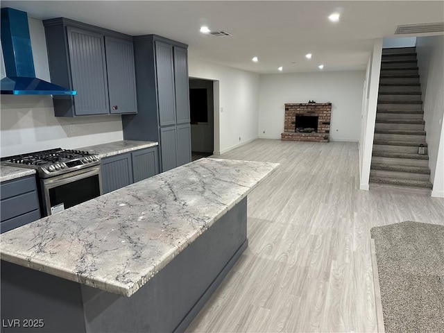 kitchen with light hardwood / wood-style flooring, stainless steel gas stove, a fireplace, a kitchen island, and wall chimney exhaust hood