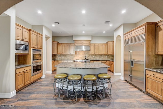 kitchen featuring backsplash, a center island with sink, built in appliances, light stone counters, and a kitchen bar