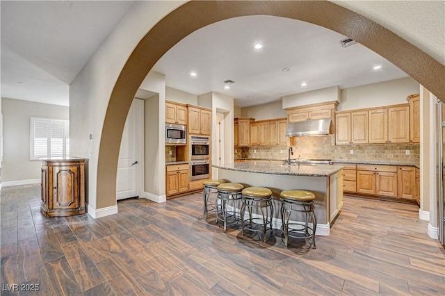 kitchen featuring dark hardwood / wood-style floors, light stone counters, stainless steel appliances, a center island with sink, and light brown cabinets